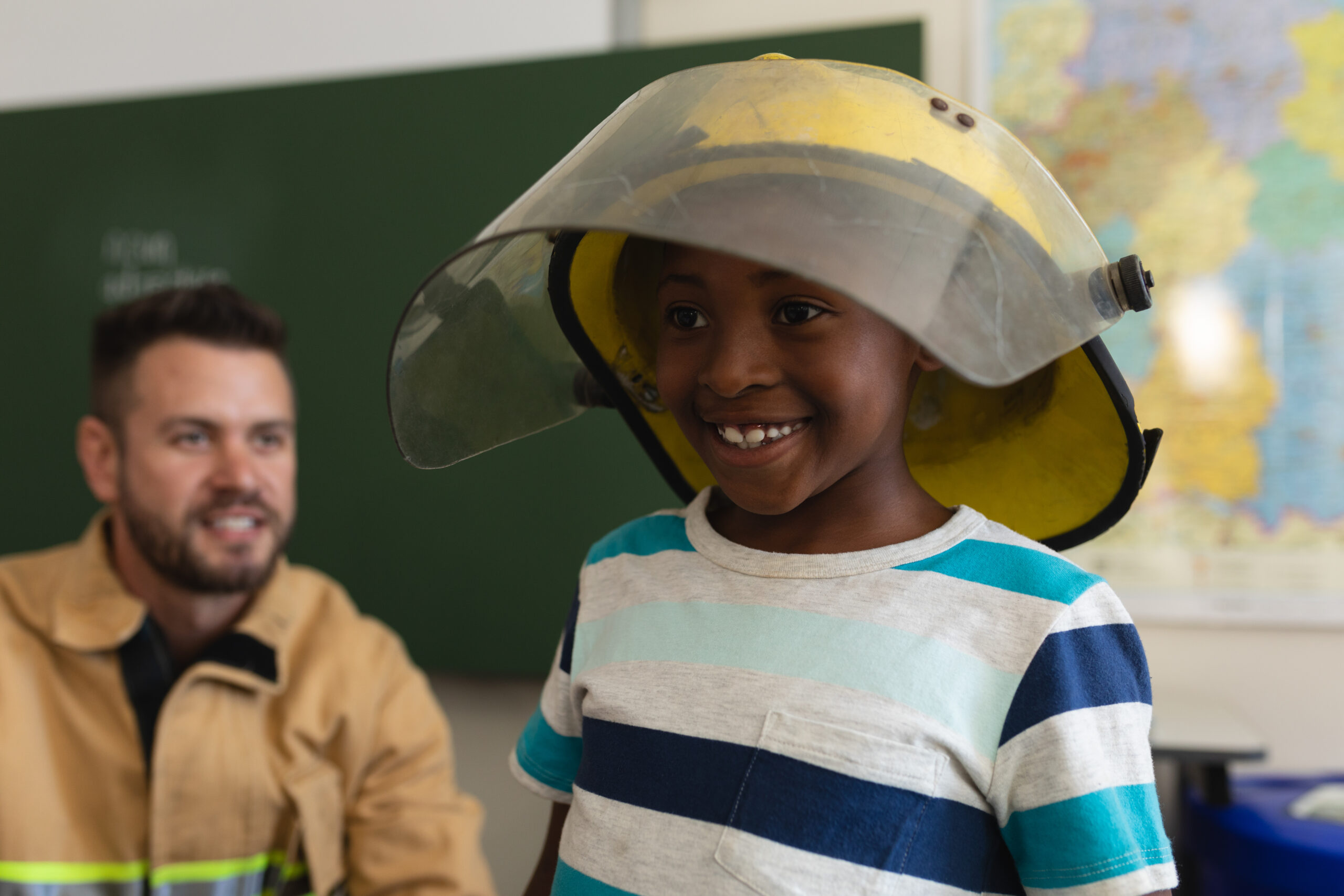 Front view of a happy black schoolboy wearing fire helmet and a smiling firefight behind him in classroom of elementary school